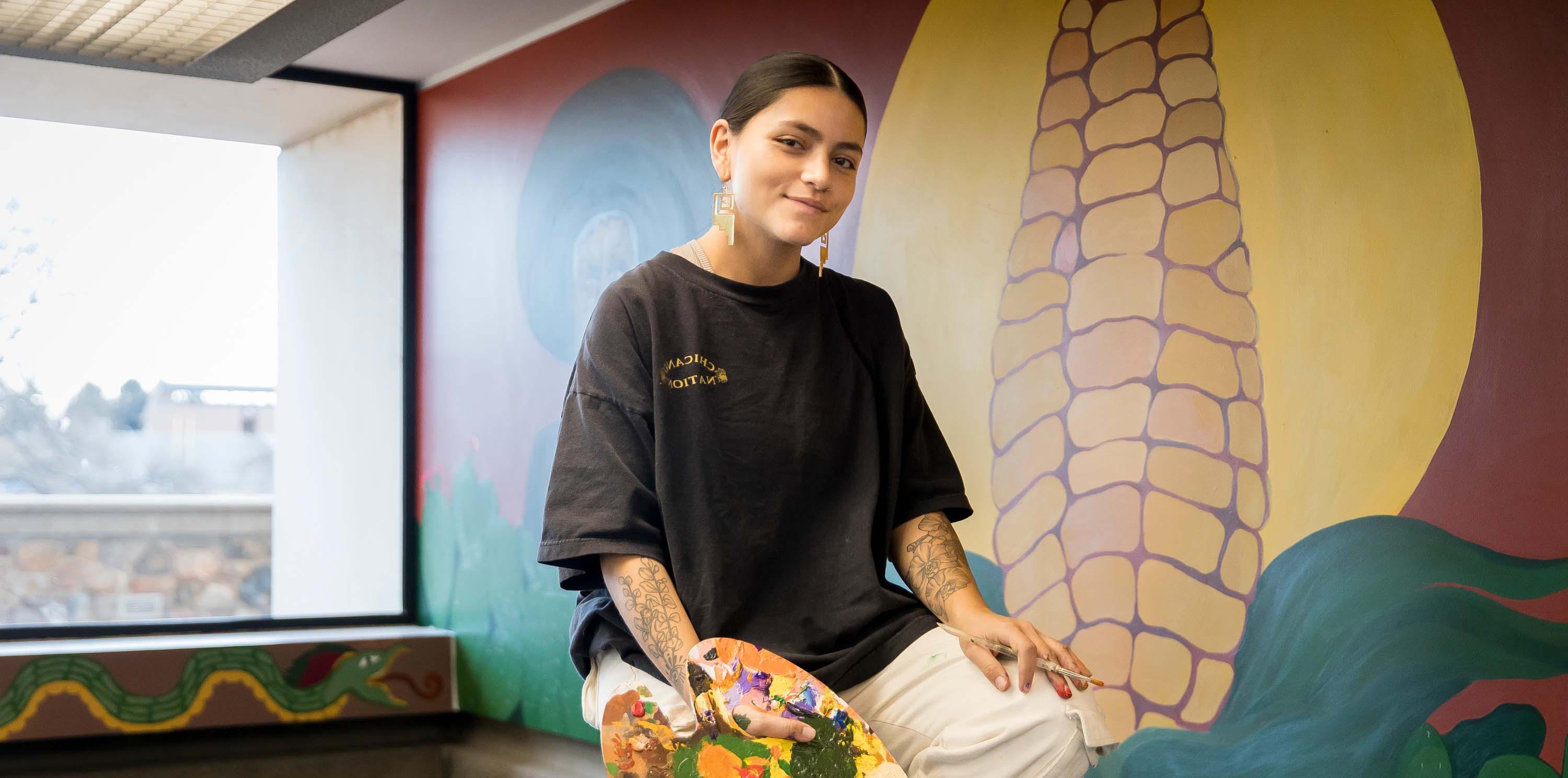 La alumna Brenda Vargas sentada en una escalera con un pincel frente a su mural