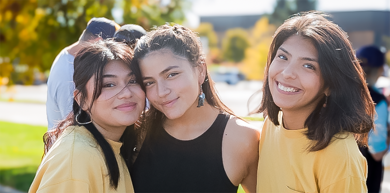 Tres sonrientes estudiantes 大学联盟 permanecen juntas y de pie en el exterior durante un acto para celebrar las comunidades hispanas/latinas. 