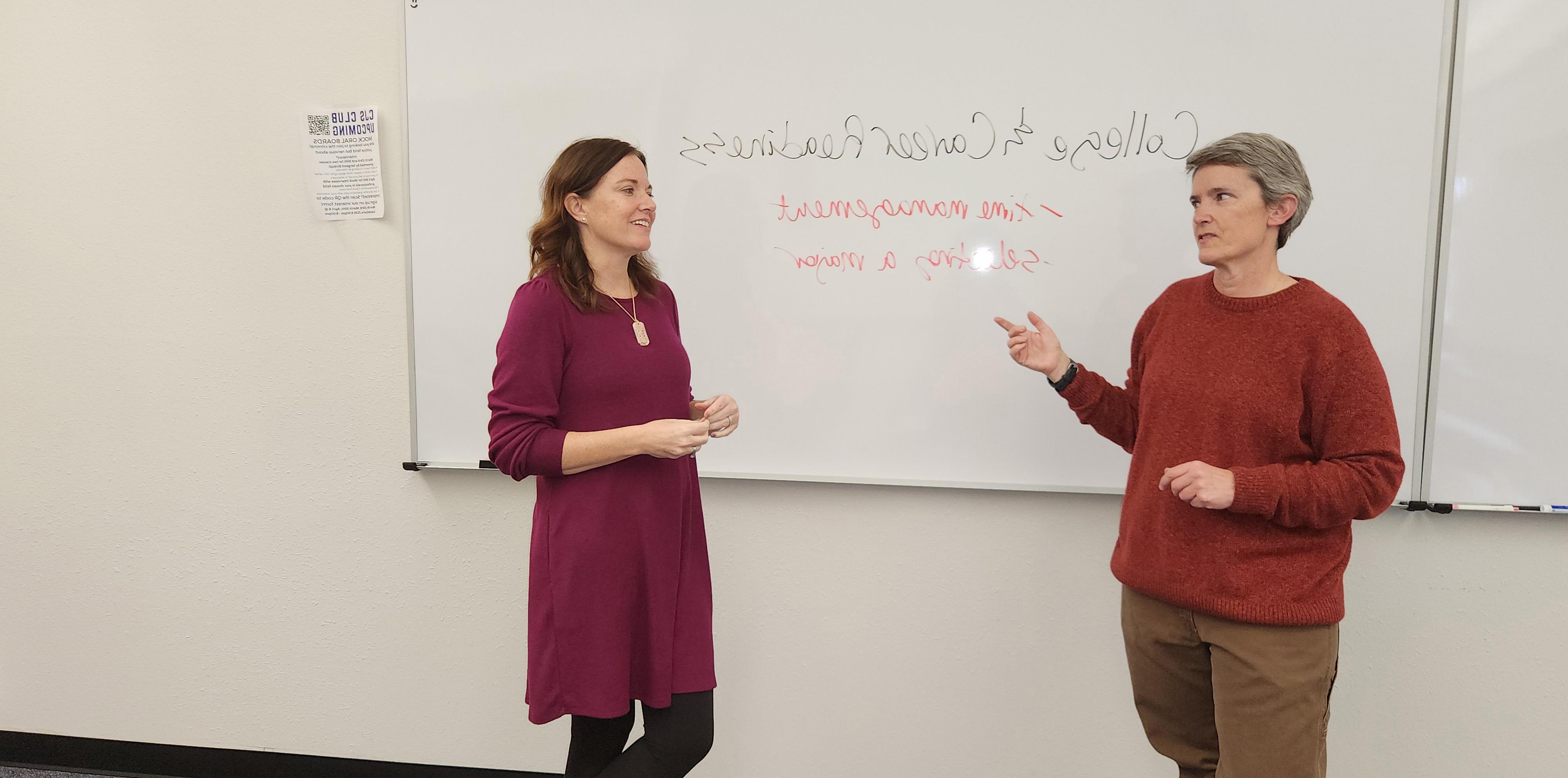 Two woman standing in front of a dry erase board looking at each other while talking