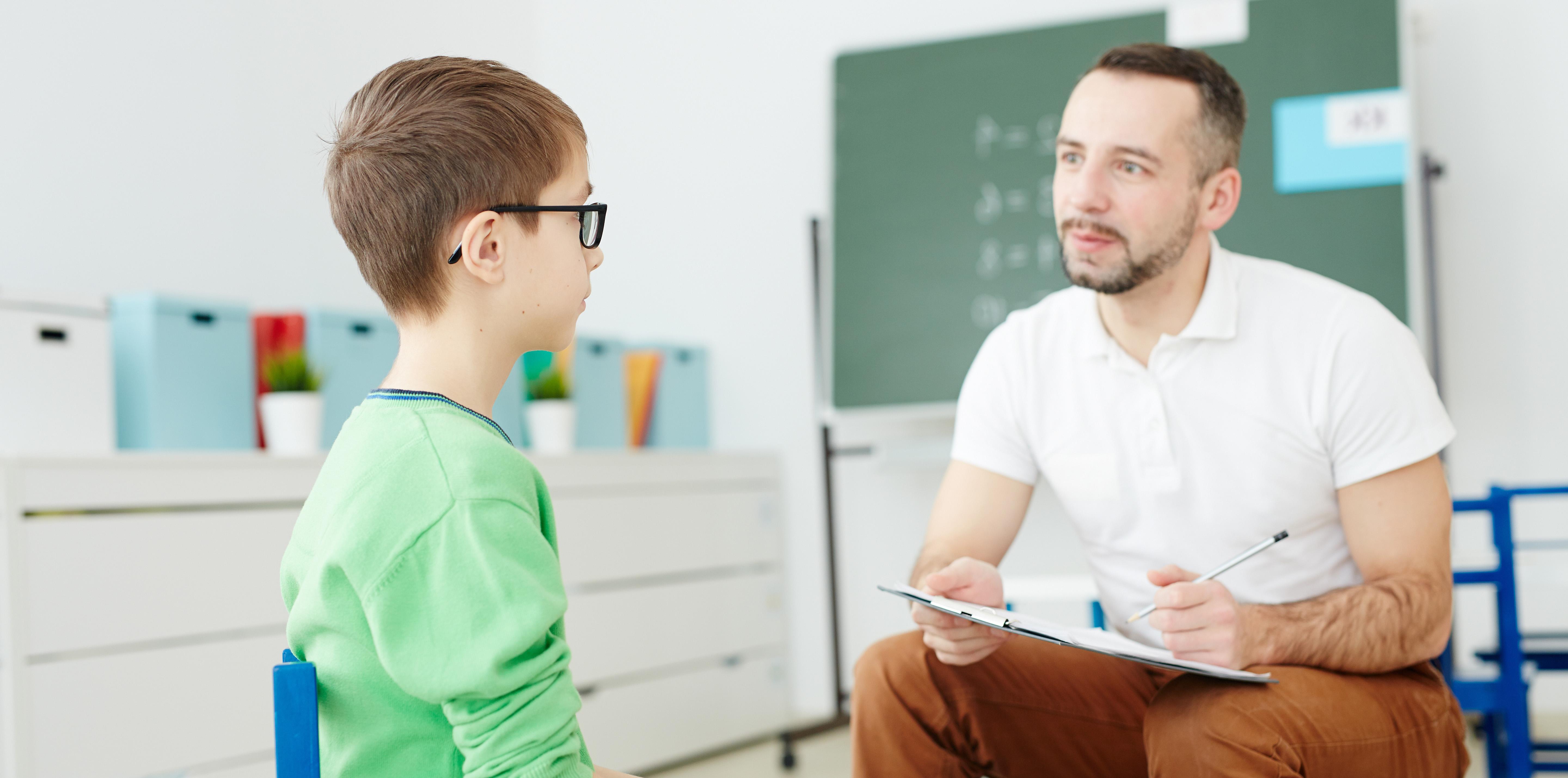 A male educator holding a clipboard sitting in a chair next to a young male student