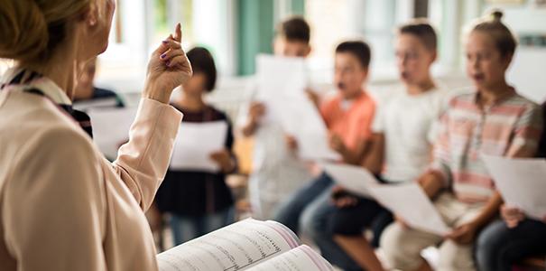 Teacher standing with music book open in front of young school-age children singing